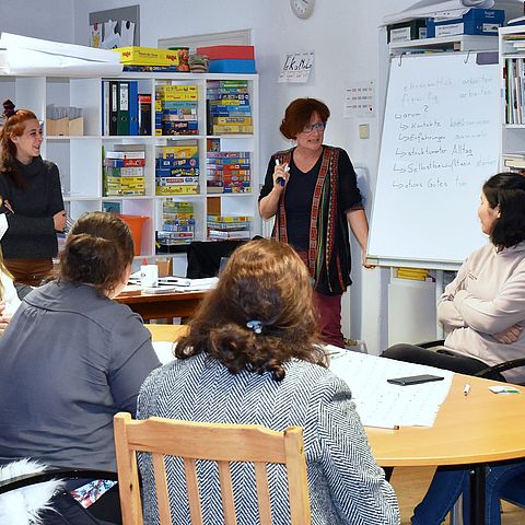 Die Teilnehmerinnen des Deutschkurses sitzen an einem großen runden Tisch; die Kursleiterin steht an der Tafel