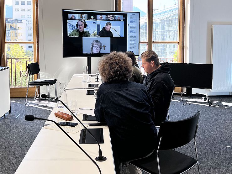 VertreterInnen sitzen im Medienraum, Bildschirm zeigt andere Online-TeilnehmerInnen