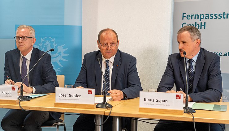 Foto bei Pressekonferenz; drei Männer in Anzügen sitzend; vor ihnen Mikrofone