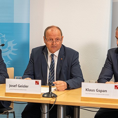 Foto bei Pressekonferenz; drei Männer in Anzügen sitzend; vor ihnen Mikrofone