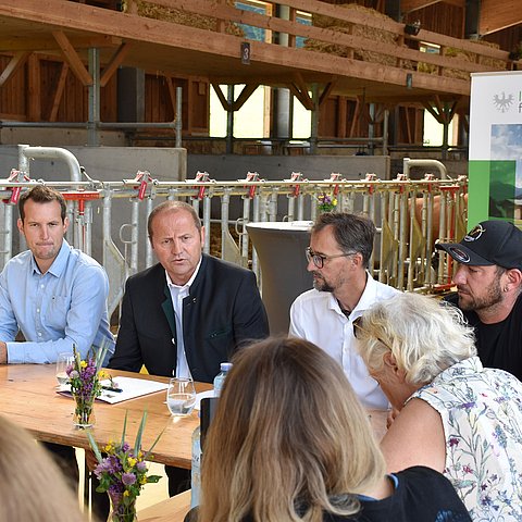 LHStv Geisler und weitere Personen sitzen an Holztisch in Kuhstall