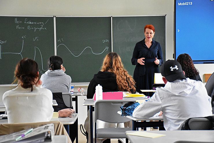 Hagele in Schulklasse stehend (vor Tafel), zu SchülerInnen sprechend