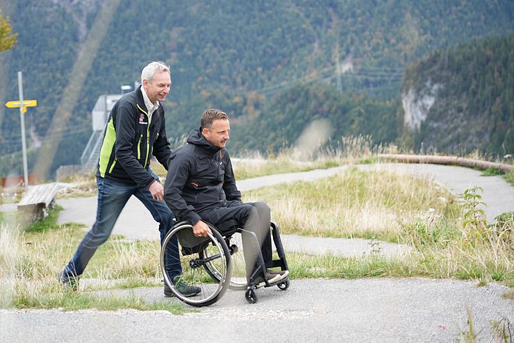 Mann schiebt Mann in Rollstuhl auf Wanderweg