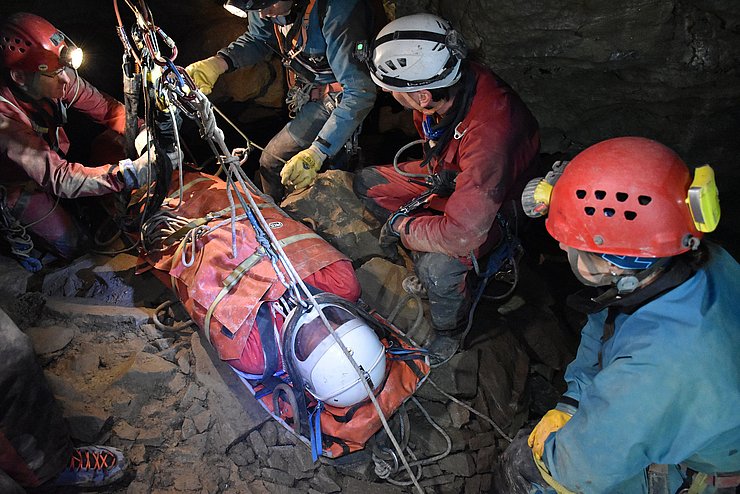 © Land Tirol/Pölzl In Höhle: Person in Trage, HelferInnen mit Helmen und Stirnlampen vergurten Trage