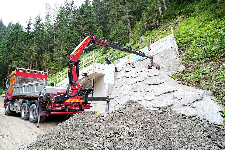 Laster mit Kranarm bei Rohbau, davor ein Haufen Erde