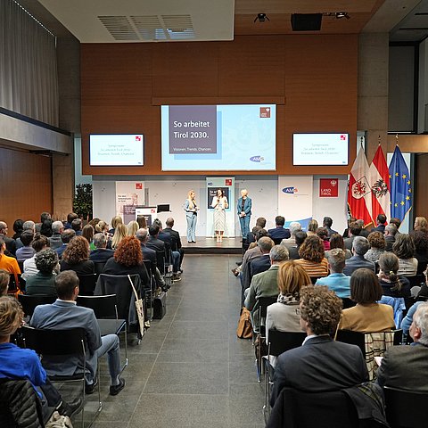 Großer Saal im Landhaus: Menschen auf Stühlen sitzend; auf Bühne drei Frauen mit Mikrofon sprechend