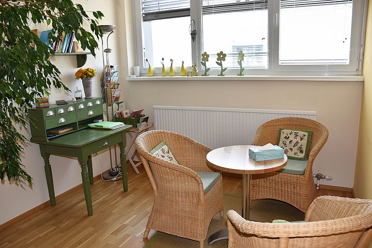 Beratungsraum mit Tisch und Stühlen aus Bast, dahinter Fenster, links ein grüner Schreibtisch