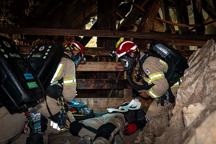 Dunkler Stollen: zwei Mitglieder der GRubenwehr mit Helm und Atemschutzmaske knieen über einer am Boden liegenden Person