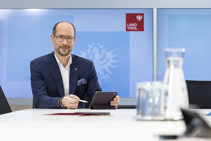 Mario Gerber sitzt mit Tablet am Schreibtisch, im Hintergrund Bildschirm mit Land Tirol Logo