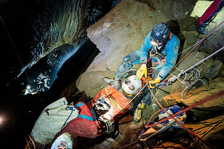 In Höhle: Person in Trage, HelferInnen mit Helmen und Stirnlampen vergurten Trage