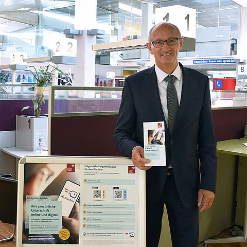 Landesrat Anton Mattle hält Informationsbroschüre zur Handy-Signatur bzw. ID Austria in der Hand, daneben ein A-Steher mit Informationsblättern