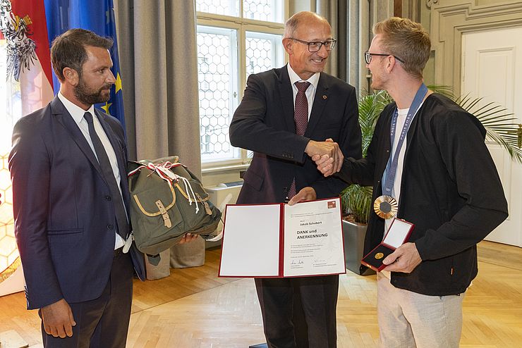 Zwei Männer überreichen einem dritten Mann eine Urkunde, eine Goldmünze und einen Rucksack als Geschenk.