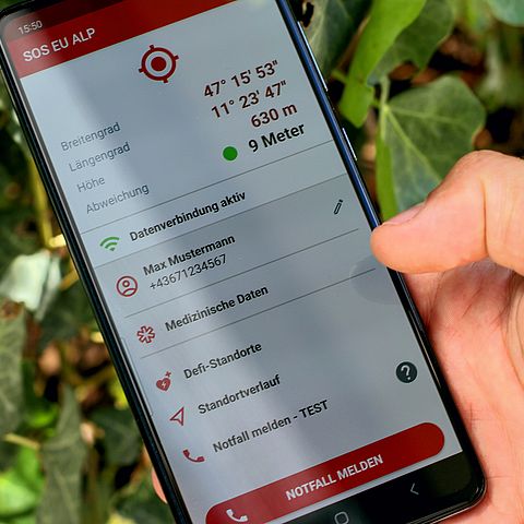 Hand hält Smartphone; Auf Smartphone ist SOS-EU-ALP-App aktiviert; zu sehen sind Koordinaten und ein großer roter Knopf mit "Notfall melden"