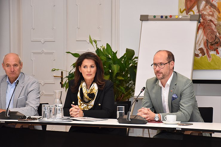 drei Personen sitzen am Tisch, eine spricht in Mikrofon