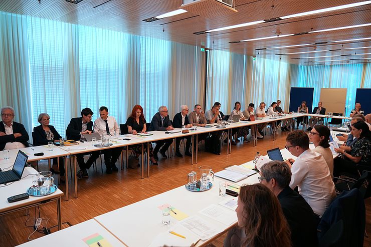 Besprechungsraum mit Tischen auf denen viele Menschen sitzen 